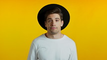 Young handsome unsure hipster guy in white shirt and hat shrugs his arms, makes gesture of I don't know, can't help anything. Yellow studio background. High quality 4k footage