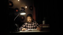 a girl sitting at a desk reading a Bible in a dark room under a lamp 