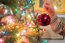A child hangs a bulb on a lit Christmas tree