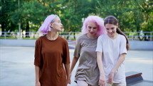 Happy teenage girls walking outdoors in green skate park. Friends having fun, laughing, holding hands. Pretty hipster women with dyed hairstyle in casual wear.


