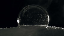 Ice crystals forming in slow motion on freezing bubble sitting on snow in front of black background.