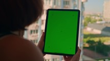 woman using digital tablet with isolated green screen tapping on display standing near open window urban city view on the background