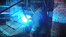 Unrecognizable man in protective clothing and mask at construction factory is engaged in welding work. Concept of building and work with metal. Sparks from modern machine