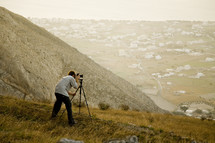 A man snaps a photo using a tripod on a hillside.