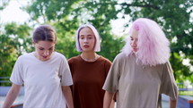Happy teenage girls walking outdoors in green skate park. Friends having fun, laughing, holding hands. Pretty hipster women with dyed hairstyle in casual wear.