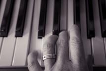 Black and white (b&w) shot of a man's hand (with wedding ring) playing a chord on a piano  keyboard