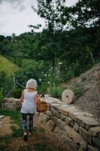 An older woman walking alongside a stone wall while carrying a basket.
