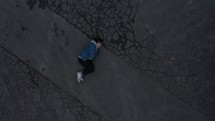 Man lays in fetal position on cracked concrete as camera slowly moves away from above