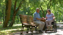 Senior man talking with wife in wheelchair outside in summer park. 