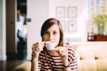 A woman sits on a couch drinking coffee from a white coffee cup.
