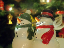 A pair of snow people snow men smile as they take in the Christmas lights of a lighted Christmas tree surrounded by CHristmas lights to celebrate the Spirit of the Christmas holidays. 