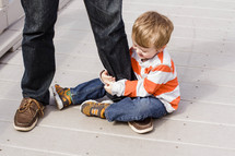 A little boy sits on the ground holding his father's leg.