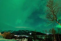 time-lapse of the northern lights with clouds moving over a mountain and lights in a city at night 