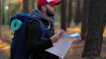 Hiking. Handsome bearded male tourist with backpack looking on map in forest. Lost hiker male with backpacks reading travel map, navigating and looking for directions in pine forest. Trekking concept.