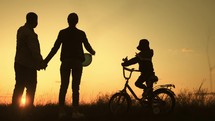 People silhouettes on summer sunset meadow. The kid is learning to ride a bike. Time to be together. Silhouette of happy family on a beautiful sunset meadow.