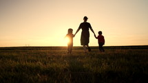 Silhouette of young family - mom and two brothers twins boys walking together to sun on open air field or park, golden hour, flares Happy mother and children, love, freedom, future concept. 4k footage