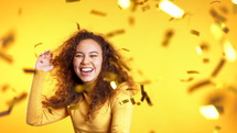 Excited girl dancing, applauding, having fun, rejoices over confetti rain in yellow studio. Concept of Christmas, New Year, happiness, party, winning.