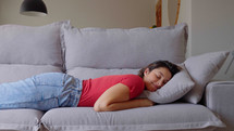 A calm young woman is resting on a couch with her eyes closed, her hands behind her head. A beautiful woman is peacefully napping on a comfortable sofa at home.
