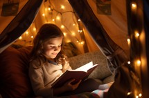 Girl engrossed in a book inside a tent, using a flashlight for illumination