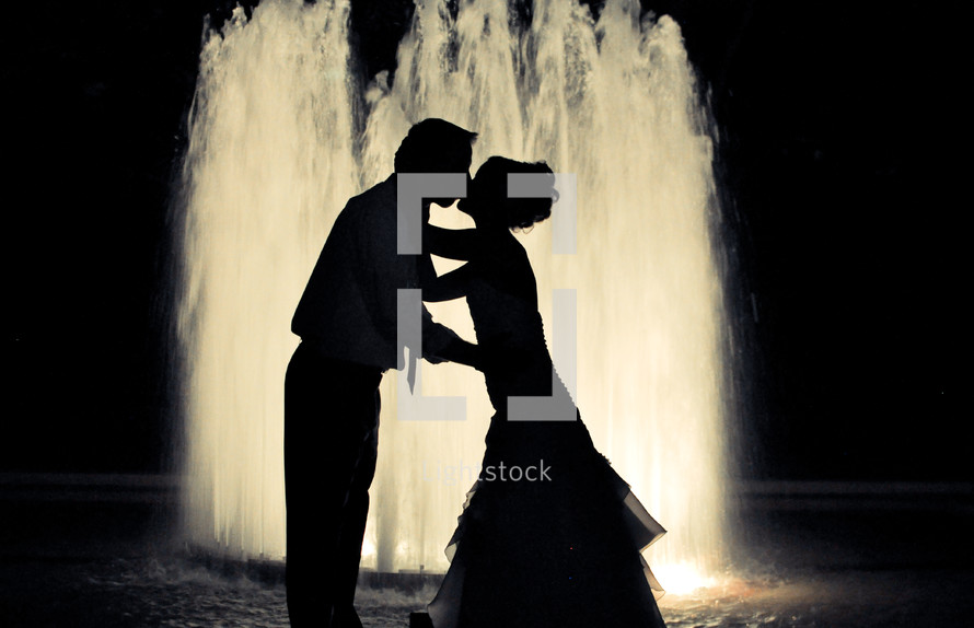 Newlyweds stand in front of a fountain kissing.