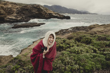 Laughing woman in red cape and head scarf holding a camera whiel standing on an oceanside hill.