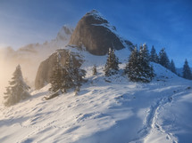 Beautiful mountain peaks with snow in winter at sunset. Colorful landscape with high, snowy rocks in morning fog, blue sky, and warm sunrise light.