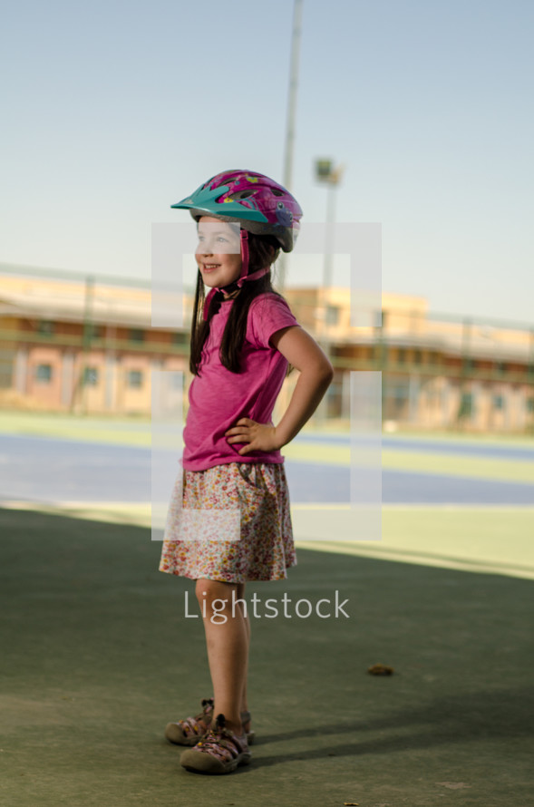 A young girl in a bicycle helmet standing near a school.