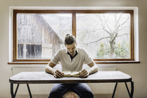 a man sitting at a desk reading a Bible with a farm scene in the window behind him 