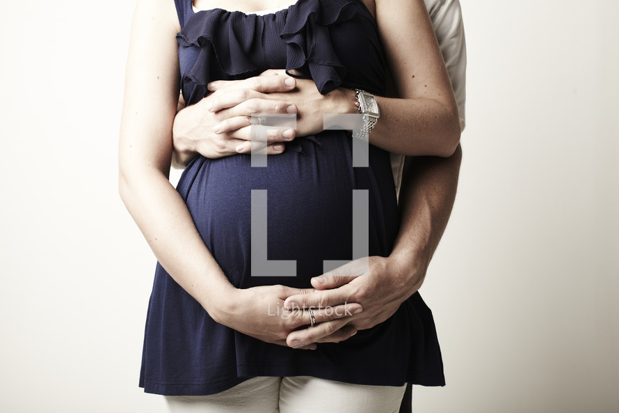 A husband and wife highlighting the fact that they are pregnant by using their interlocked hands to showcase her baby bump.   
