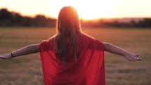 Girl Plays Superhero. Kid run across green field in red cloak at sunset time. Pretty child superhero hero in red cloak in nature. Girl power concept.
