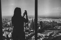 A woman takes a picture of a city from a high rise window.