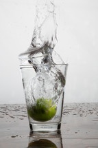 Water splashing out of a glass as a lime is dropped into the glass.