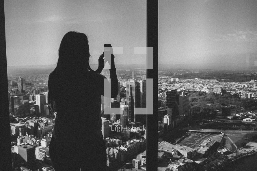 A woman takes a picture of a city from a high rise window.