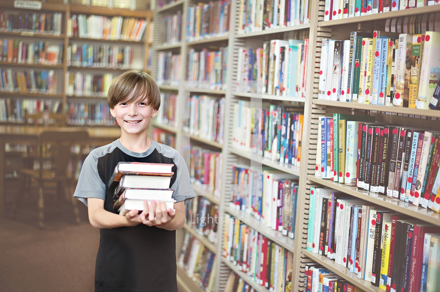 Smiling boy carrying a stack of books in a library.