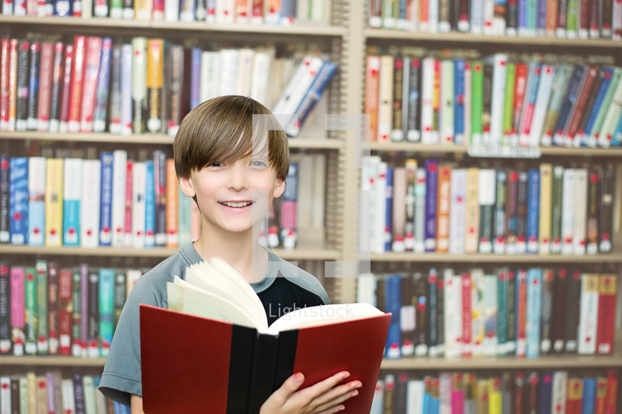 Smiling boy holding an open book in a library.