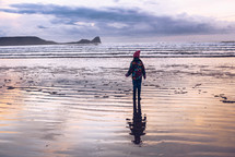 a girl in a winter coat walking on wet sand along a shore 