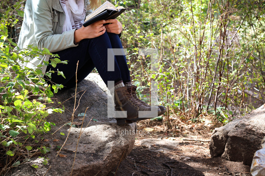 A woman sits outdoors on a rock and reads her Bible.