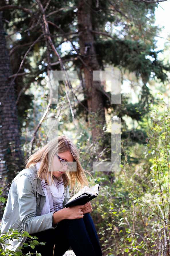 A woman sits and reads her Bible in a forest.