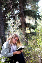 A woman sits and reads her Bible in a forest.