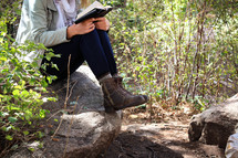 A woman sits outdoors on a rock and reads her Bible.