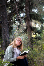 A young woman reads her Bible and thinks in a forest.