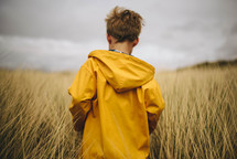 a boy in a rain coat walking through tall grass in a field 