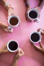 The hands of four women holding coffee cups at a pink table.