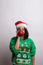 woman in an ugly Christmas sweater and santa hat hiding behind a Christmas ornament 