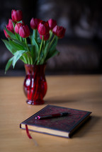 A vase of red tulips on a table next to a journal and pen.