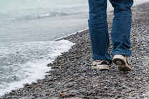 A man's legs walking on a gravel shore.