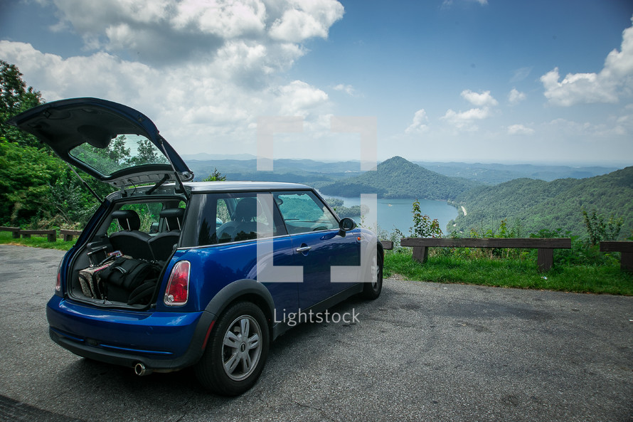 A small blue car parked at an overlook and facing mountains and a lake.