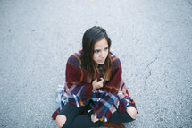 a lost teen wrapped in a blanket sitting on asphalt 