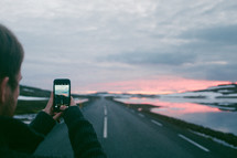 a man taking a picture with his cellphone of a rural highway at sunset 