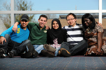 Five young people of different ethic backgrounds, arms around each other, sitting on a floor outdoors